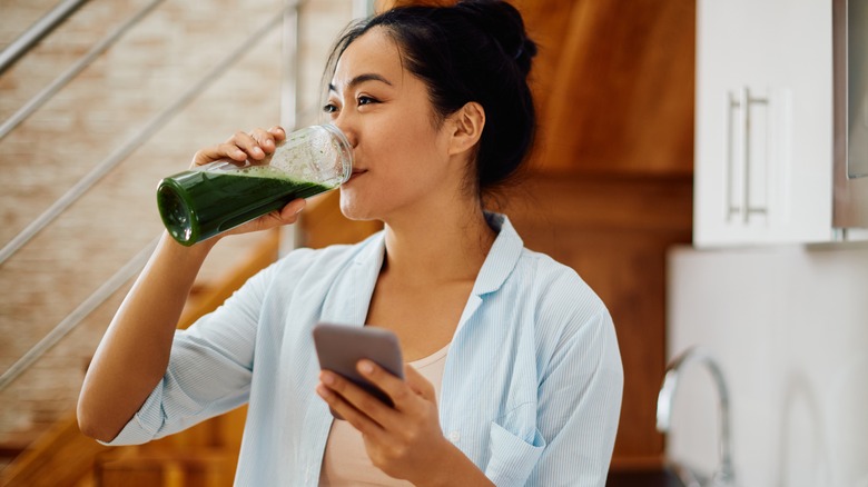 Woman drinks a green juice while holding her phone