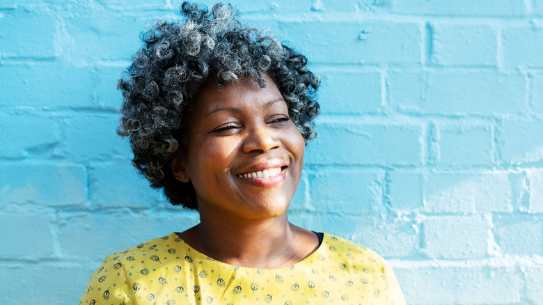 A smiling woman with a bob and wearing a yellow top standing in front of a wall.