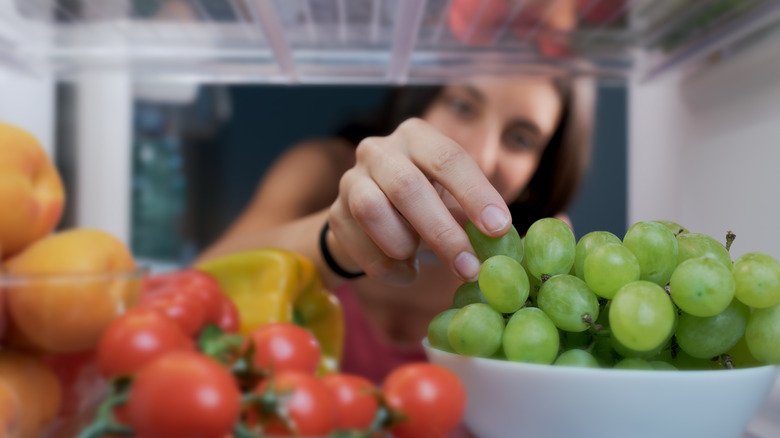 Woman reaching into fridge filled with fresh produce, including grapes and tomatoes.