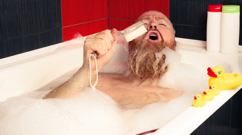 A man lying in a bubble bath using a brush as a microphone