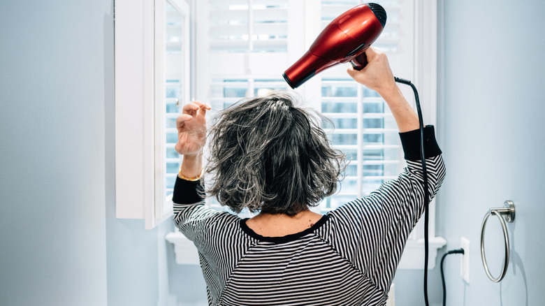 An older woman with gray hair blow drying her hair in the bathroom.