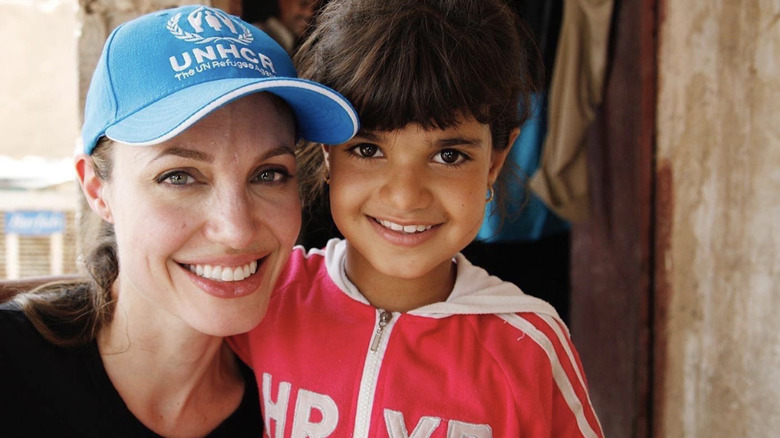 Angelina Jolie wears a cap and minimal makeup as she smiles with a small child.