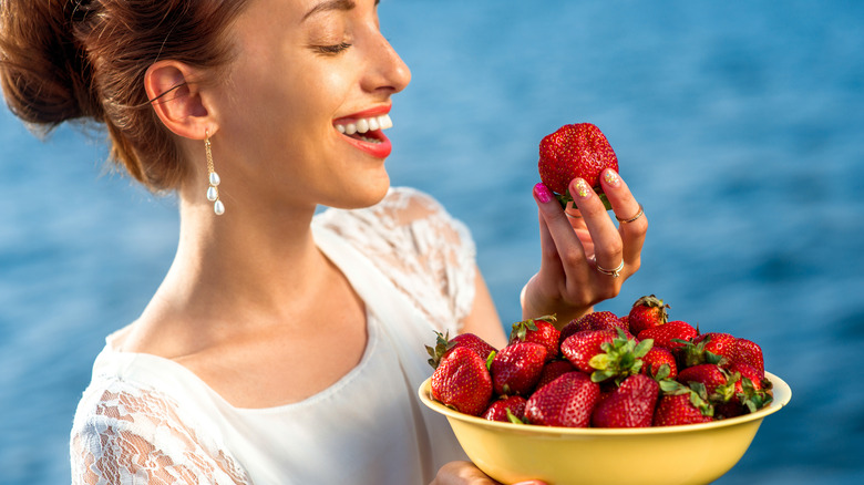 Woman holding strawberry in one hand and bowl of strawberries in other hand