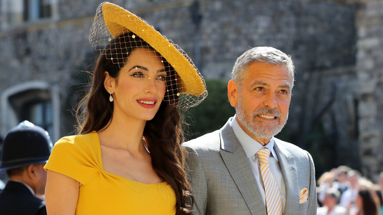 Amal Clooney, in a yellow dress and fascinator, with George Clooney