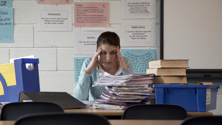 a woman sits behind a desk stacked with papers, hands on her head