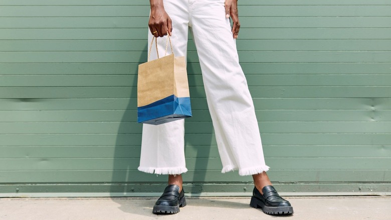 A person wearing white cropped pants with frayed cuffs and black shoes, carrying a shopping bag