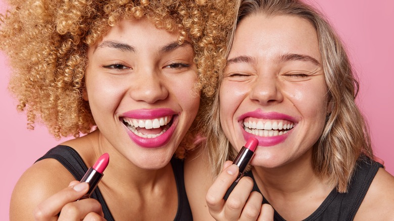 two female wearing black tops holding pink lipsticks smiling