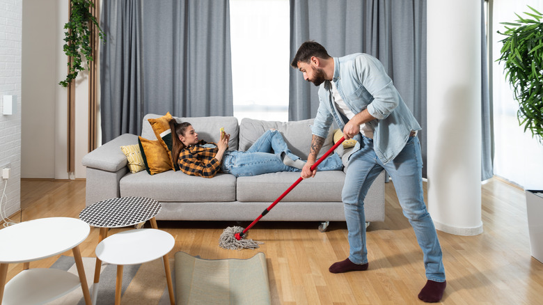 man mopping while his wife lies on the couch looking at her phone