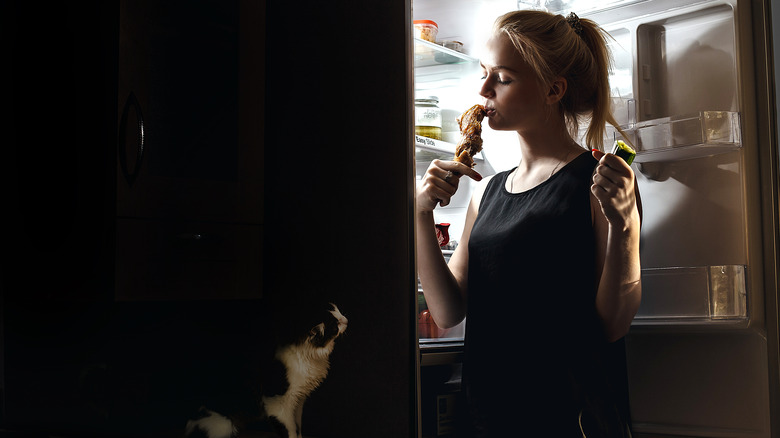 Woman eating from the fridge while cat looks on