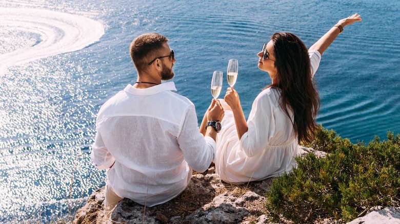 a couple sitting on a cliff facing the sea drinking champagne