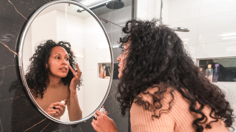 A middle-aged woman applying a product to her skin in the mirror.