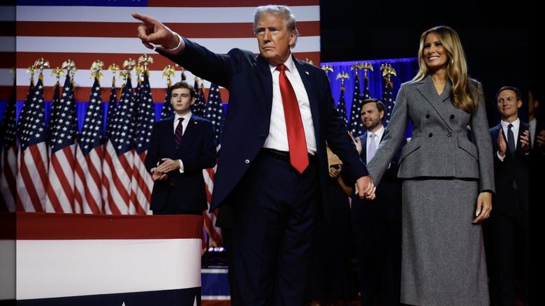 Melania Trump standing with Donald Trump on stage in a gray skirt suit.