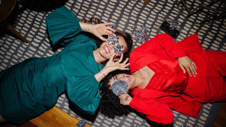 Two women lying on their backs in festive dresses holding silver holiday boys over their eyes