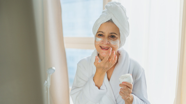 A middle-aged woman applying cream to her face while in a bathrobe