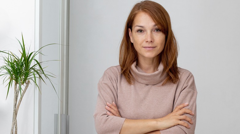 An older woman with auburn hair stands with her arms crossed.