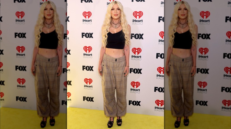 Tori Spelling in a cropped top and baggy pants in front of a media wall.