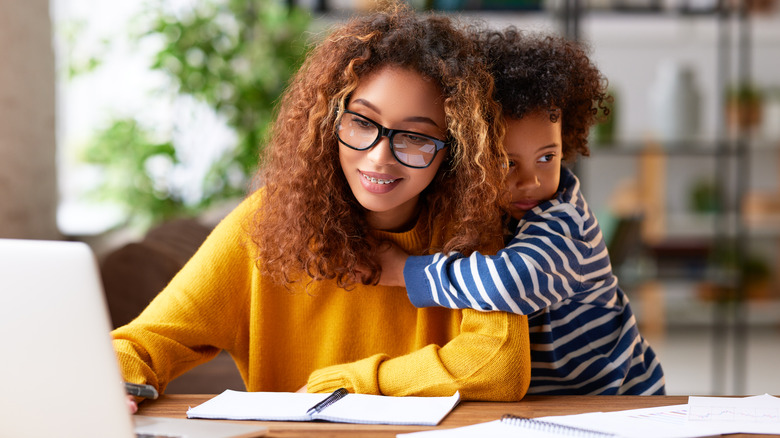 Woman works from home while child hangs onto her shoulder 