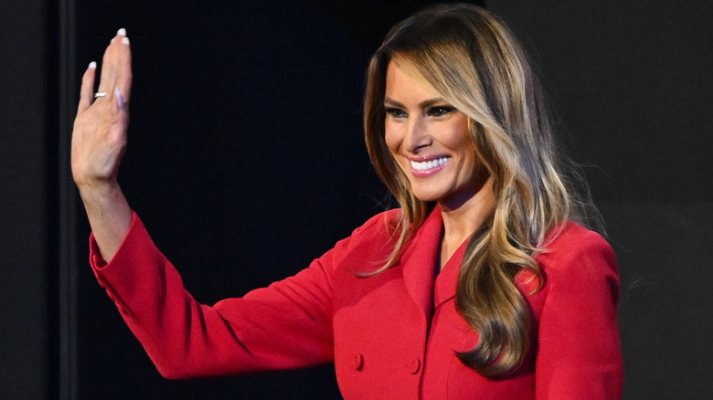 Melania Trump smiling and waving in a red blazer.