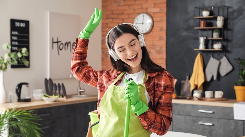 woman dances to music while wearing green cleaning gloves, apron, and holding mop