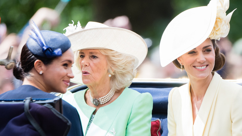 Meghan Markle, Queen Camilla, and Kate Middleton sitting in a carriage wearing colorful clothing and fancy hats